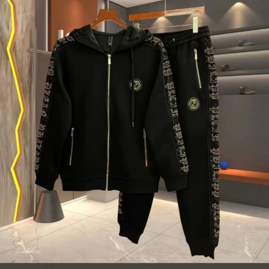 A black zip-up hoodie and matching jogger pants set hanging on wooden hangers. The hoodie has patterned black-and-white printed sleeves, a front zipper, drawstrings, and a small circular chest emblem.