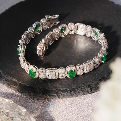 A silver-toned tennis bracelet with round and baguette-cut green emerald-cut gemstones, displayed on a dark slate slab.