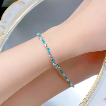 A delicate silver-toned bracelet with alternating turquoise and clear faceted gemstones, shown worn on a person's wrist against a soft, reflective background.