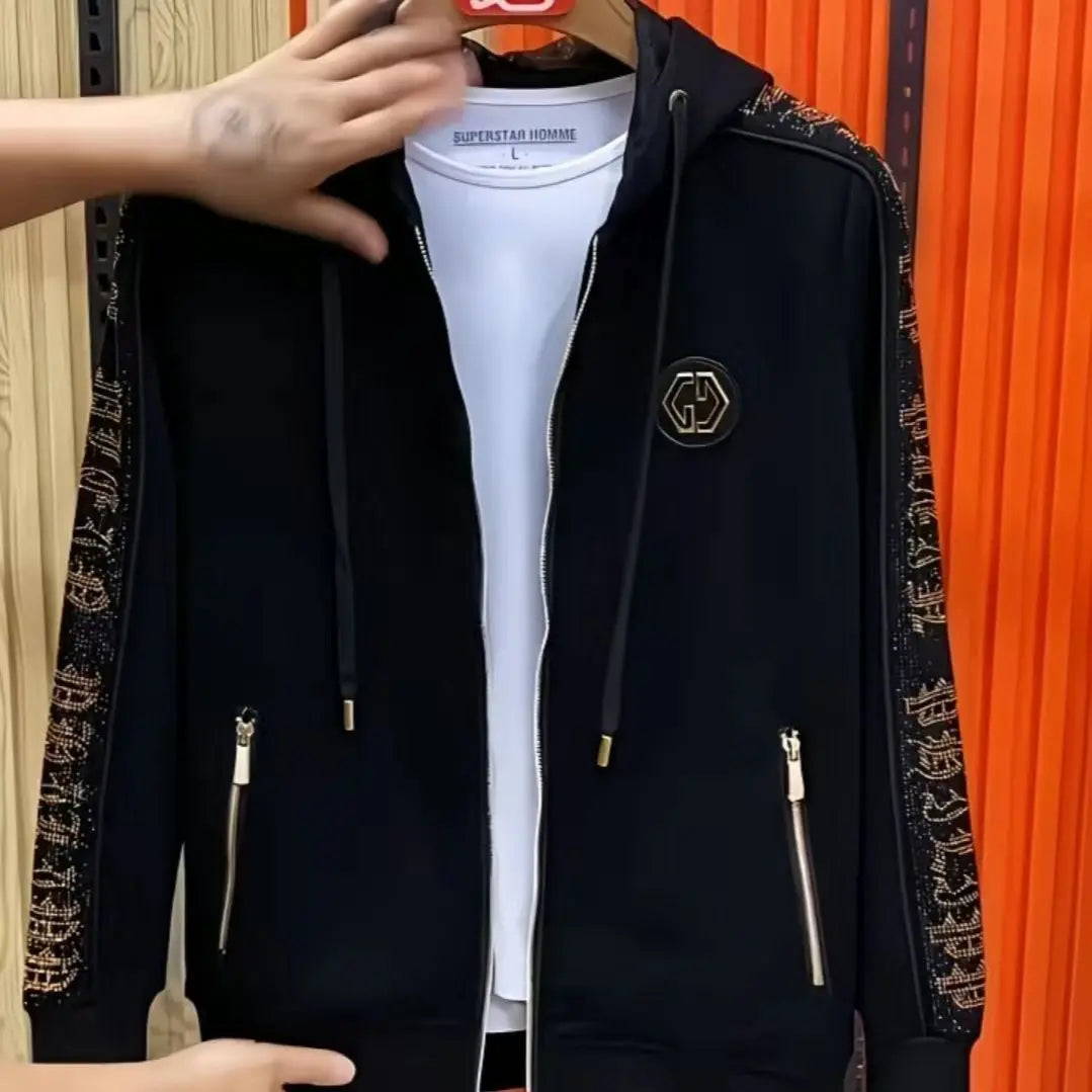 A black zip-up hoodie jacket with ornate gold patterned sleeves and a gold chest emblem, layered over a white t-shirt. The hoodie is shown hanging on a wooden hanger against a background of light wood paneling on the left and orange vertical slats on the right.