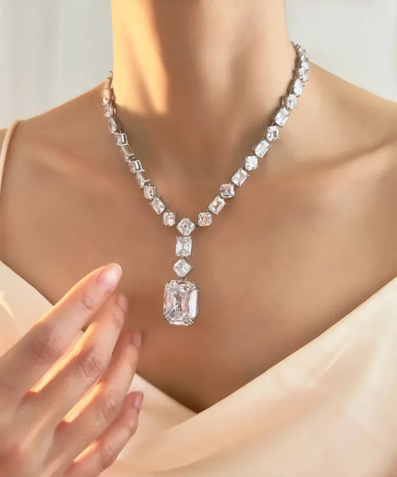 A close-up of a model wearing a sparkling silver-toned necklace with a large rectangular emerald-cut pendant and matching clear stones along the chain.