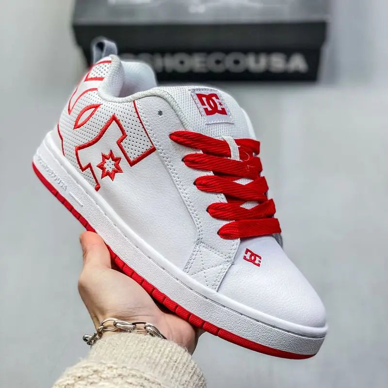 A hand holds a white DC Shoes low-top sneaker with bright red laces, red side logo details, and a red outsole, with a gray background and a shoebox visible behind it.