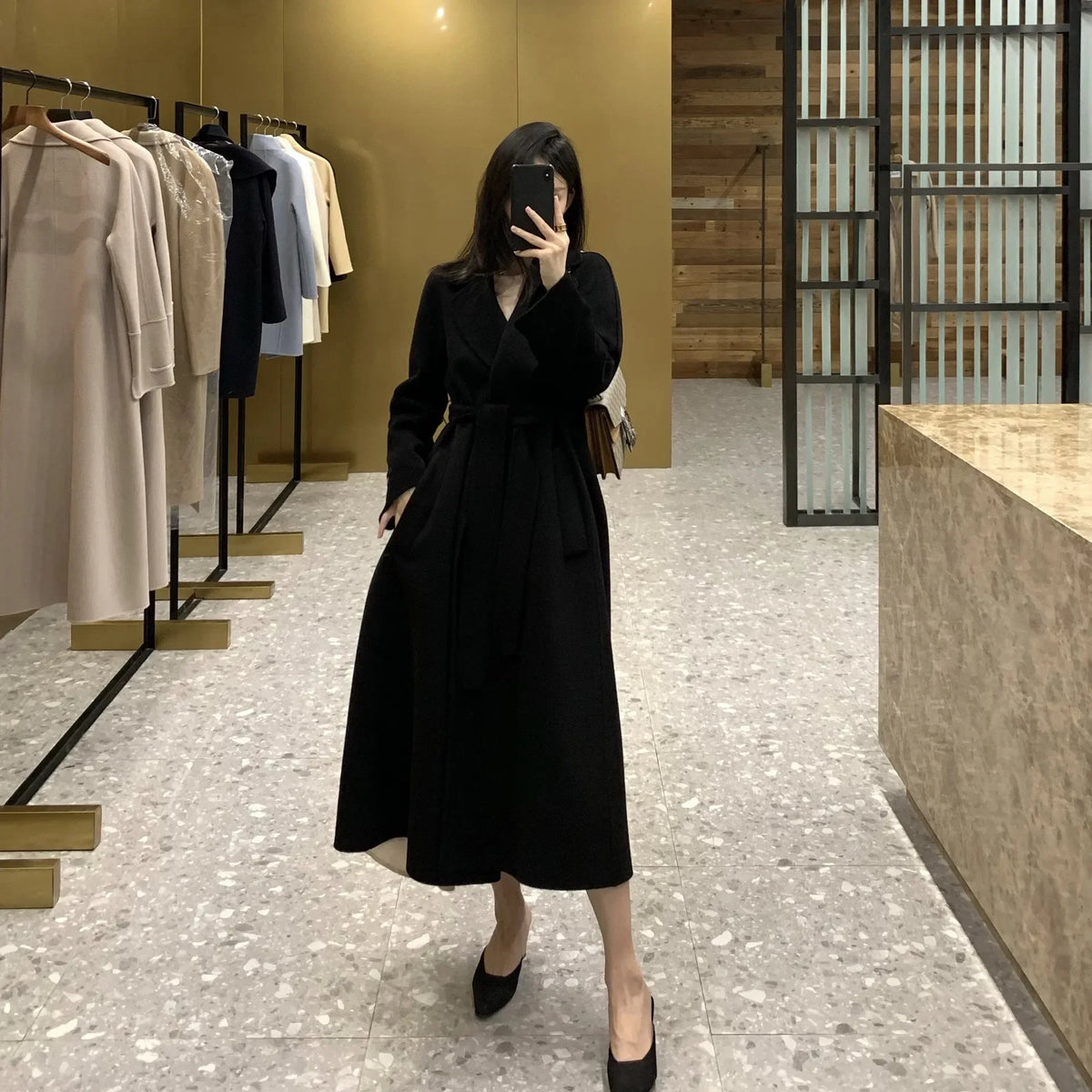 A woman takes a mirror selfie in a clothing boutique wearing a long black wrap-style midi dress with a tie waist belt. The boutique features a gold-toned wall, racks of neutral-colored shirts, and a marble-patterned floor.