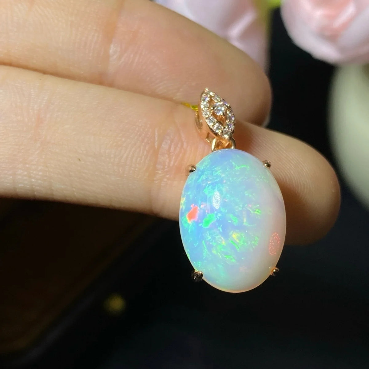 A close-up of a hand holding a gold pendant with an oval white opal stone showing vibrant blue, green, and orange flashes, accented by small clear stones in the bail.