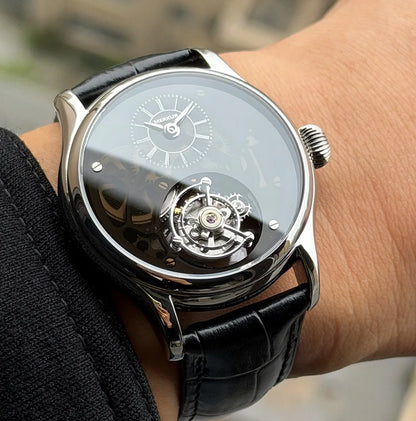 A close-up of a wrist wearing a luxury skeleton watch with a black leather strap, silver-toned case, and a black dial showing the inner mechanical movement.