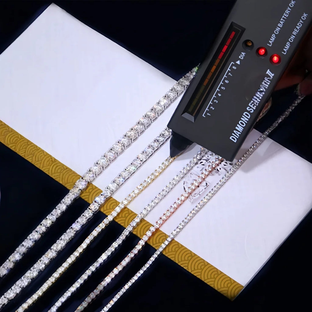 A diamond semi-quantitative tester device with red and green indicator lights and a scale, shown alongside several thin diamond-encrusted chains laid out on a white and gold card against a black background.