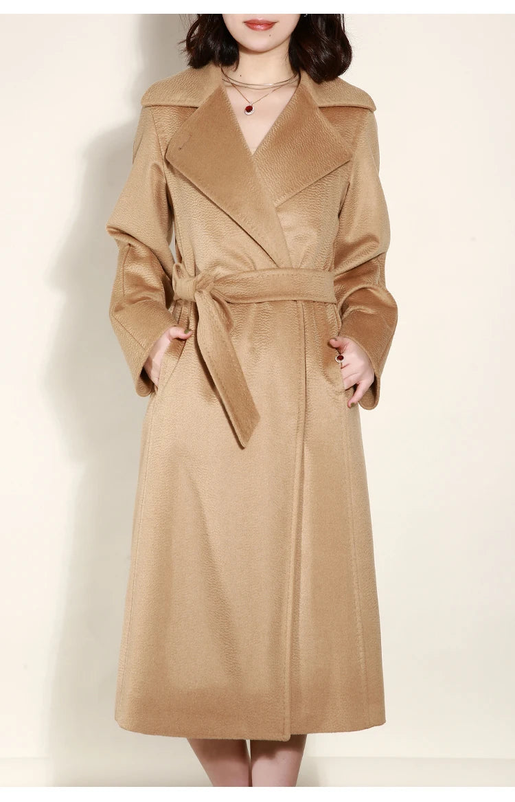 A woman wearing a long camel-colored trench coat with a self-tie belt, standing against a plain off-white background.
