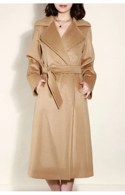 A woman wearing a long camel-colored trench coat with a self-tie belt, standing against a plain off-white background.