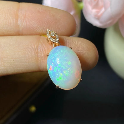 A close-up of a hand holding a gold pendant with an oval white opal stone that shows iridescent blue, green, and pink flashes, with small clear stones in the bail.