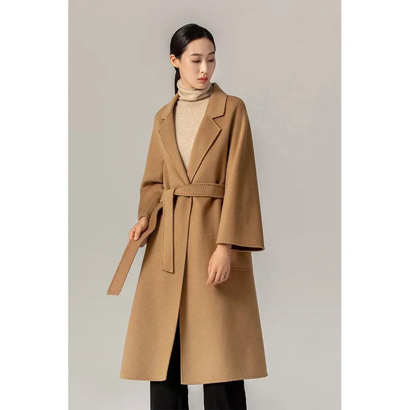 A woman modeling a camel brown belted coat over a beige turtleneck sweater and black pants, standing against a neutral light gray studio background.