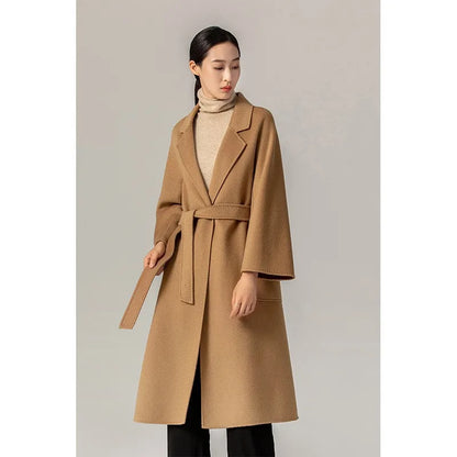 A woman modeling a camel brown belted coat over a beige turtleneck sweater and black pants, standing against a neutral light gray studio background.