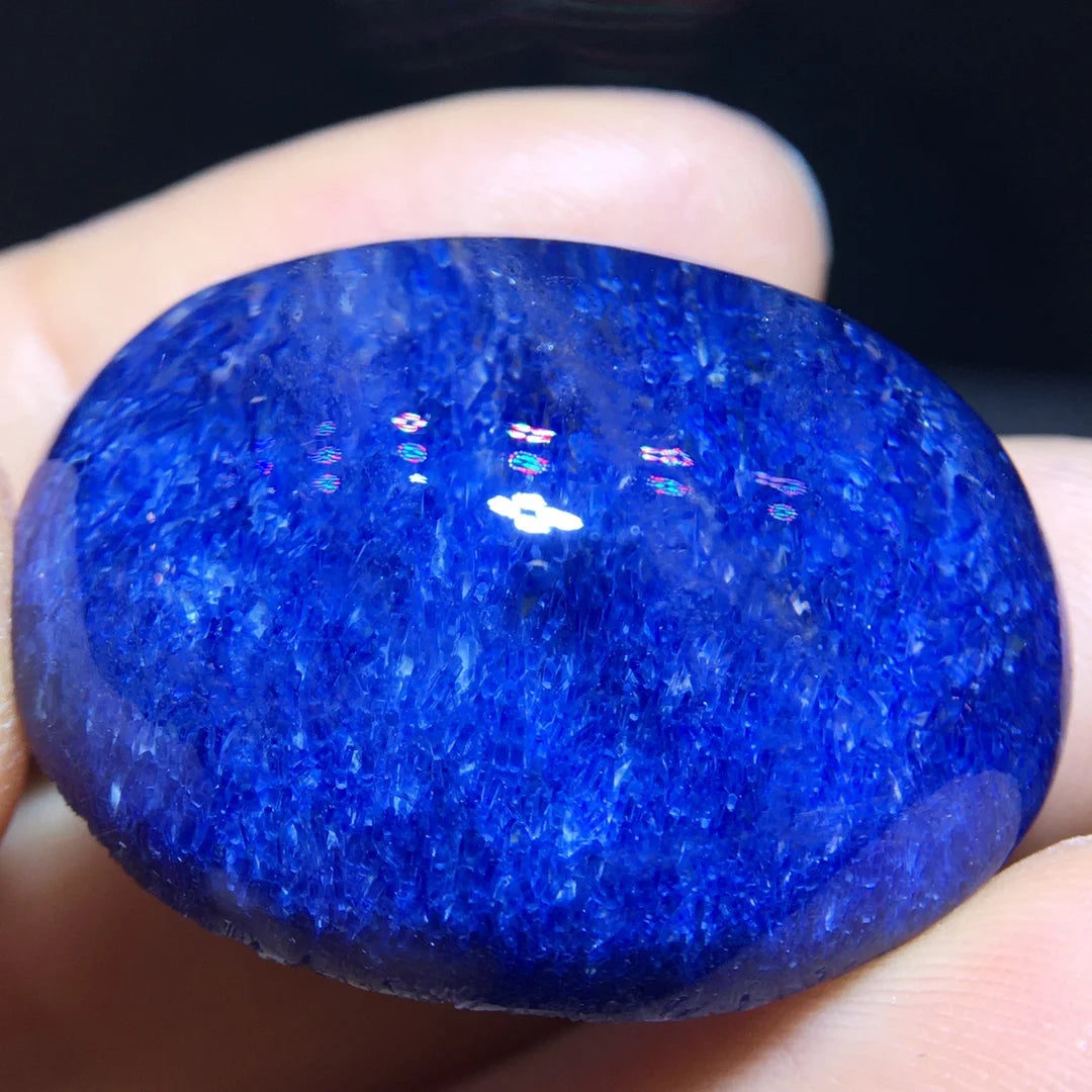 A polished oval-shaped cobalt blue stone, likely a tanzanite or sapphire gemstone, held in a person's fingers against a dark background.