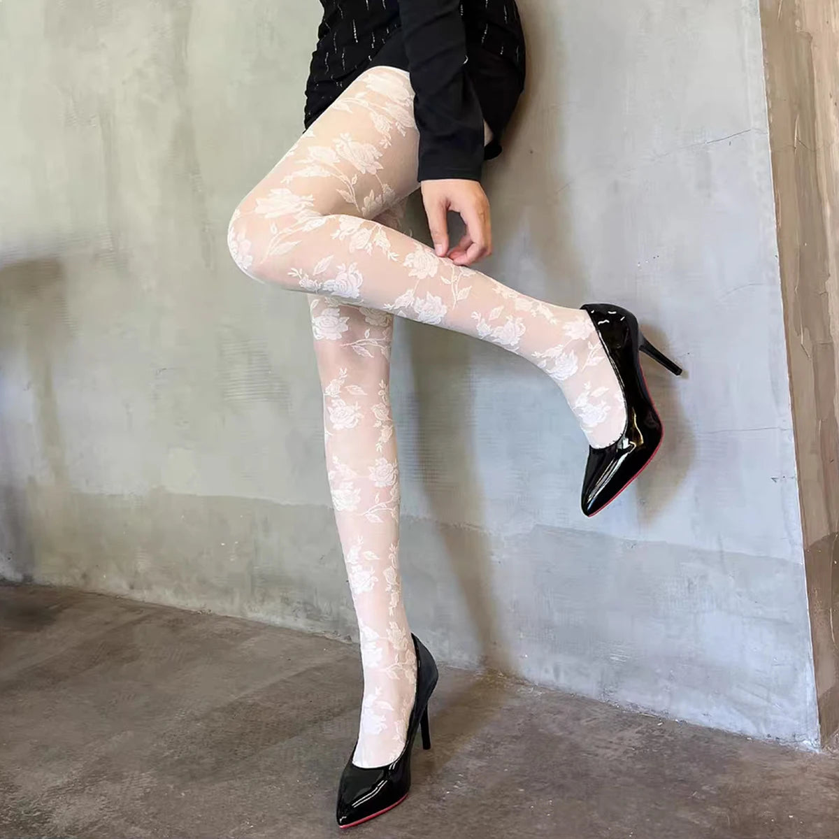 A model's legs wearing sheer beige floral lace tights and black patent leather high heels with red soles, posing against a textured concrete wall.