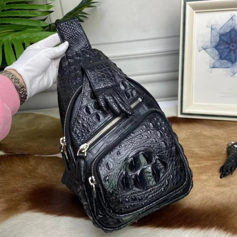 A black crocodile-embossed leather sling bag with multiple zippered compartments and a top handle, held by a white-gloved hand over a cowhide surface with a framed print in the background.