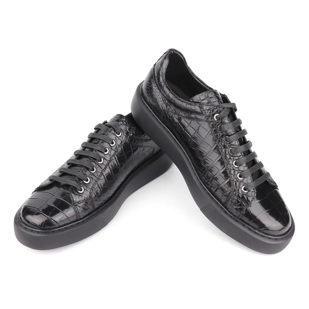 A pair of black low-top sneakers with a glossy crocodile-embossed texture, black laces, and chunky black soles, displayed on a clean white background.