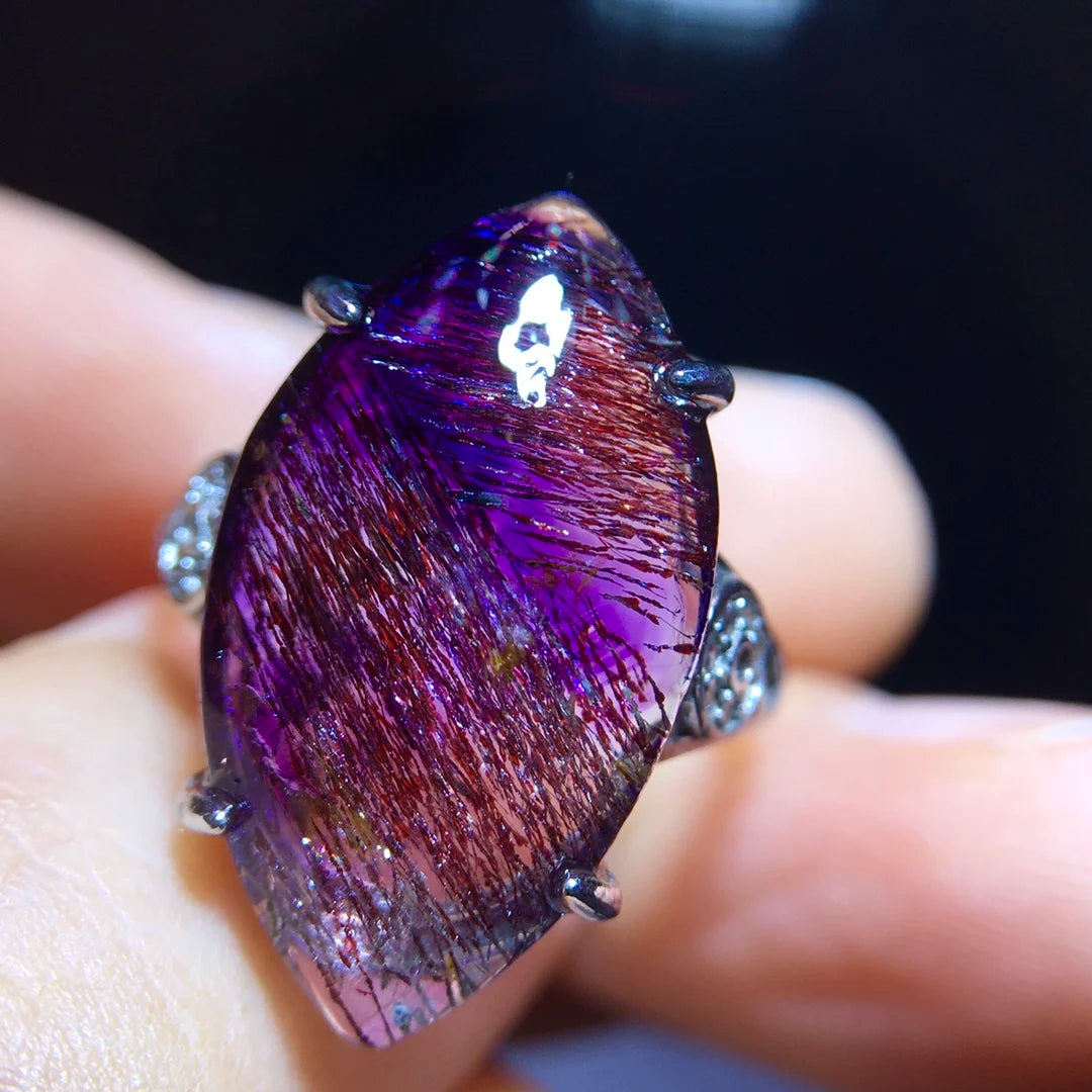 A close-up of a ring held on fingers, featuring a large teardrop-shaped purple iridescent gemstone with visible metallic needle-like inclusions and silver-toned prongs.