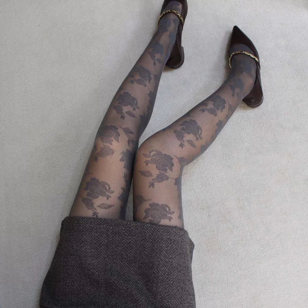 A person's legs wearing sheer black floral tights, paired with dark brown pointed-toe flats with gold chain details and a gray skirt, photographed from above on a light gray carpet.