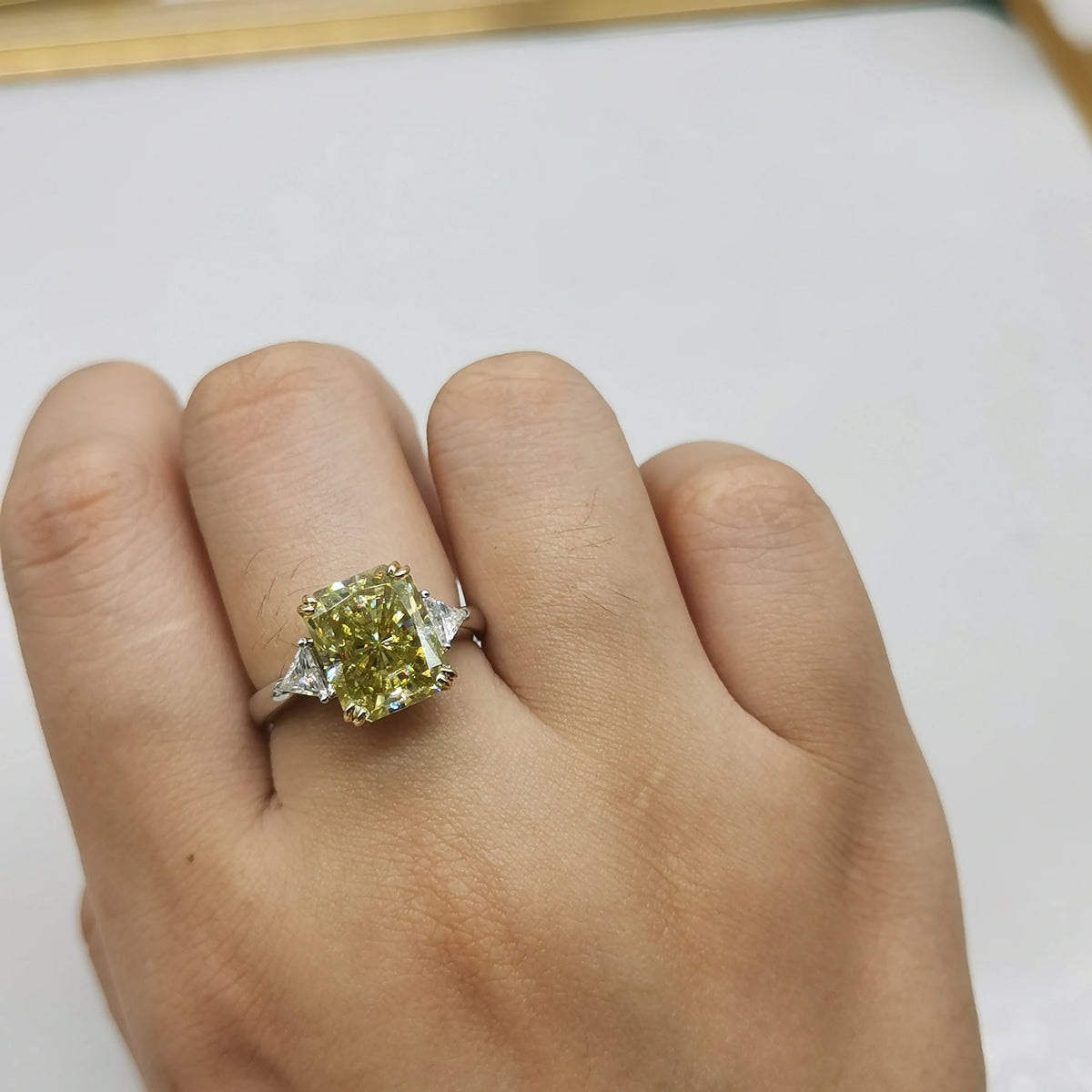A close-up of a hand wearing a ring with a large cushion-cut yellow gemstone and small side stones on a silver-toned band, set against a plain light background.