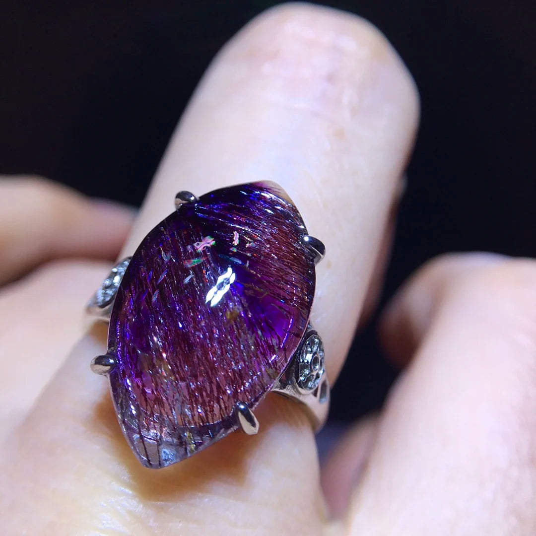 A close-up of a silver ring with a large teardrop-shaped purple iridescent gemstone held by four prongs, worn on a finger against a dark background.