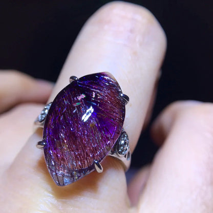 A close-up of a silver ring with a large teardrop-shaped purple iridescent gemstone held by four prongs, worn on a finger against a dark background.