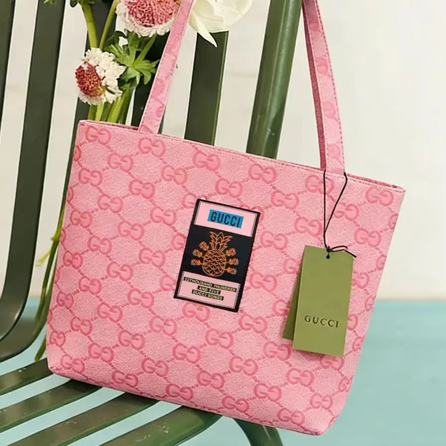 Pink Gucci tote bag with an allover GG monogram pattern, a central patch, and a green Gucci hangtag, displayed on a green chair with flowers in the background.