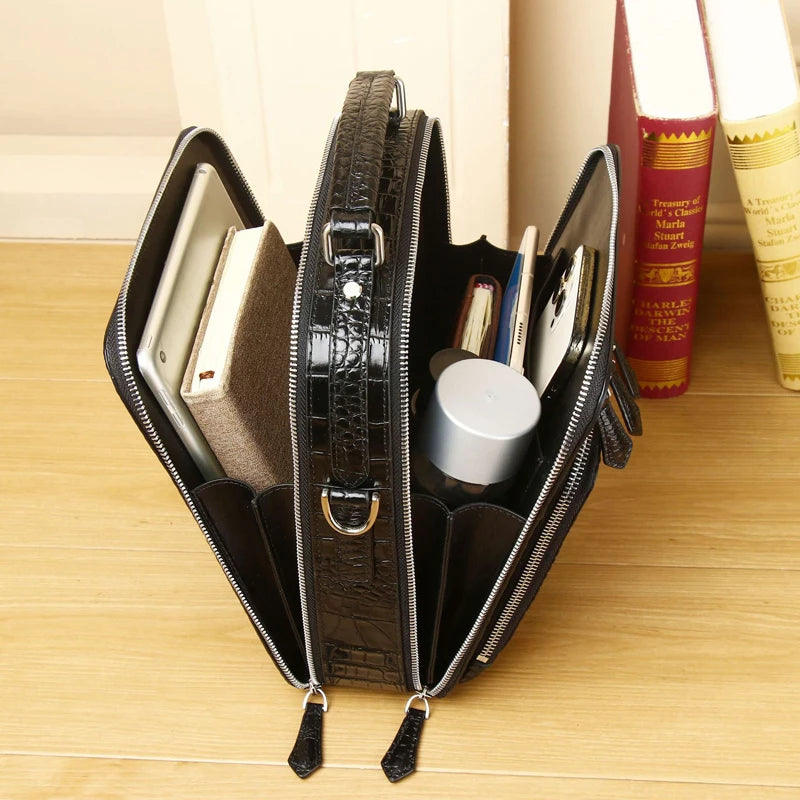 A black crocodile-embossed zip-around organizer bag shown open on a light wood tabletop, holding notebooks, a small bottle, and other accessories, with books in the background.