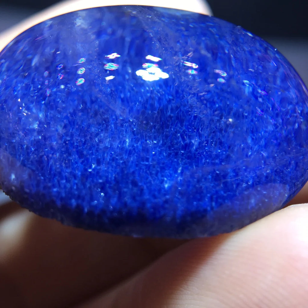 A close-up of a polished oval blue stone or gemstone held against fingers, showing a glossy, highly textured deep blue surface with bright reflective highlights.