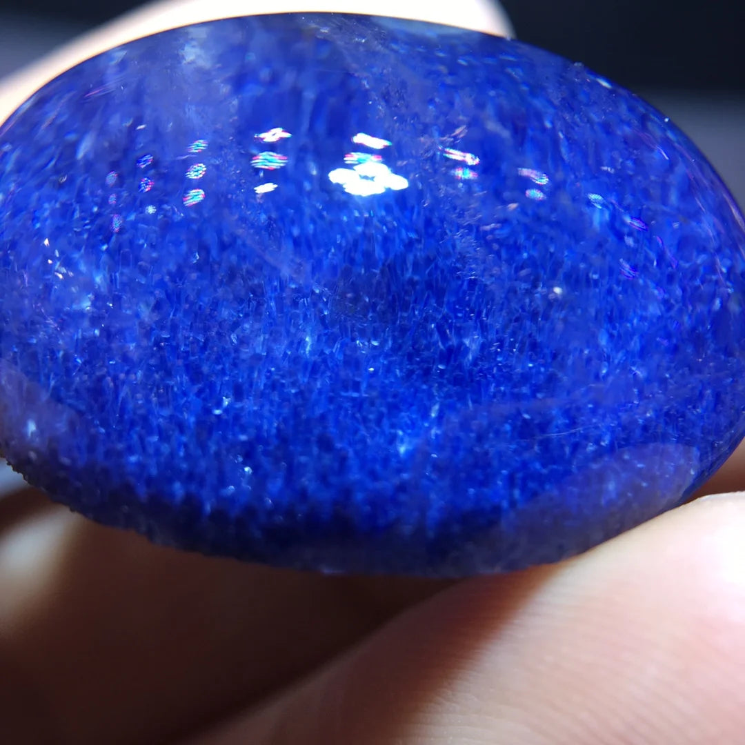 A close-up of a polished oval blue stone or gemstone held against fingers, showing a glossy, highly textured deep blue surface with bright reflective highlights.