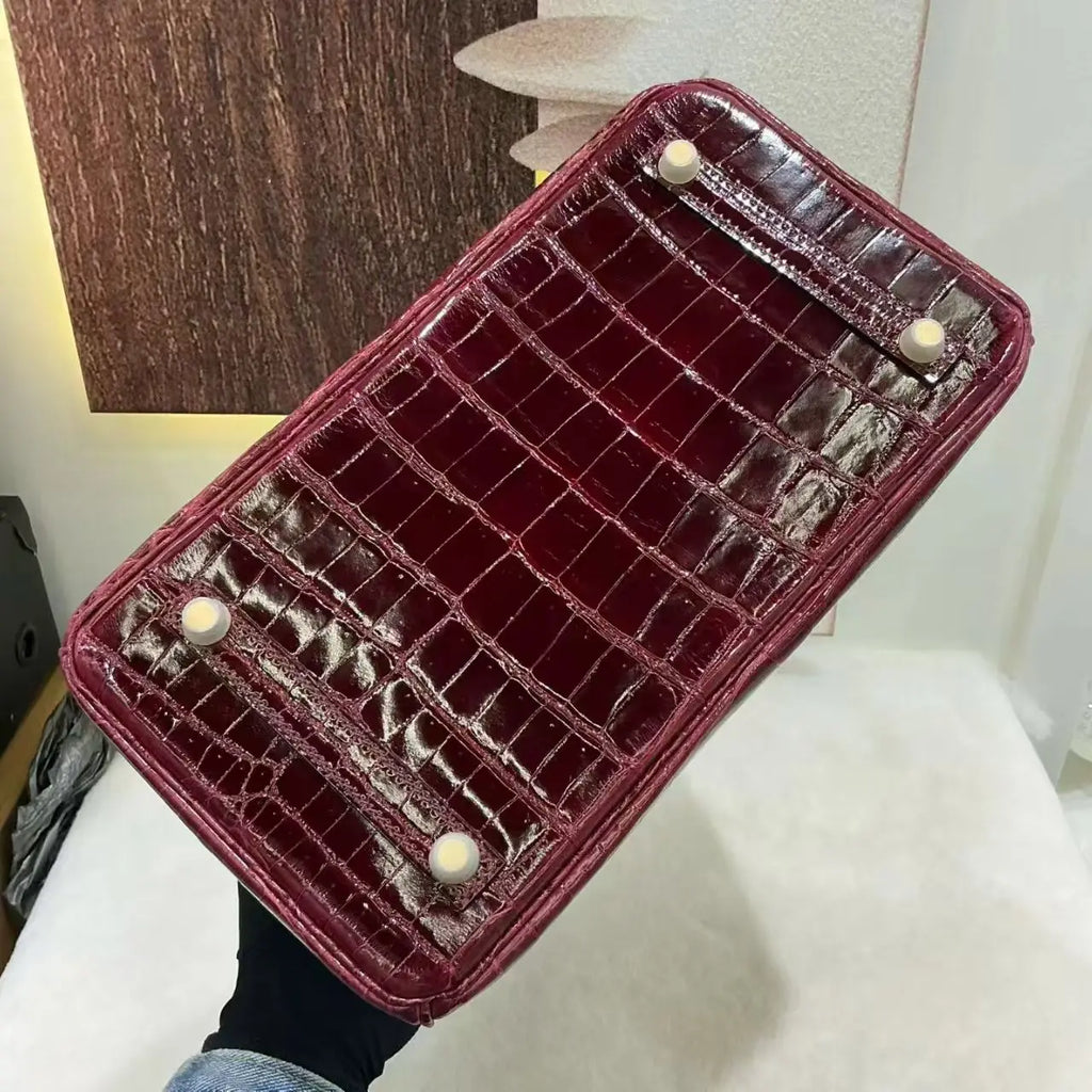 A glossy crocodile-embossed leather handbag shown from the bottom, held by a hand. The bag is a deep burgundy red with four cream-colored round studs on the bottom corners.
