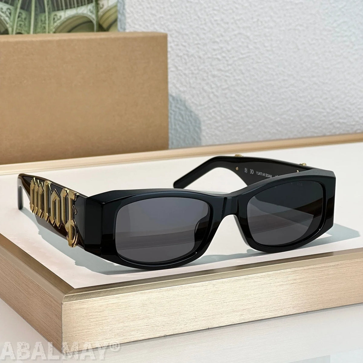 Black square sunglasses with dark lenses and ornate gold-tone decorative arm details, displayed on a light wood riser with a textured wall and box in the background.