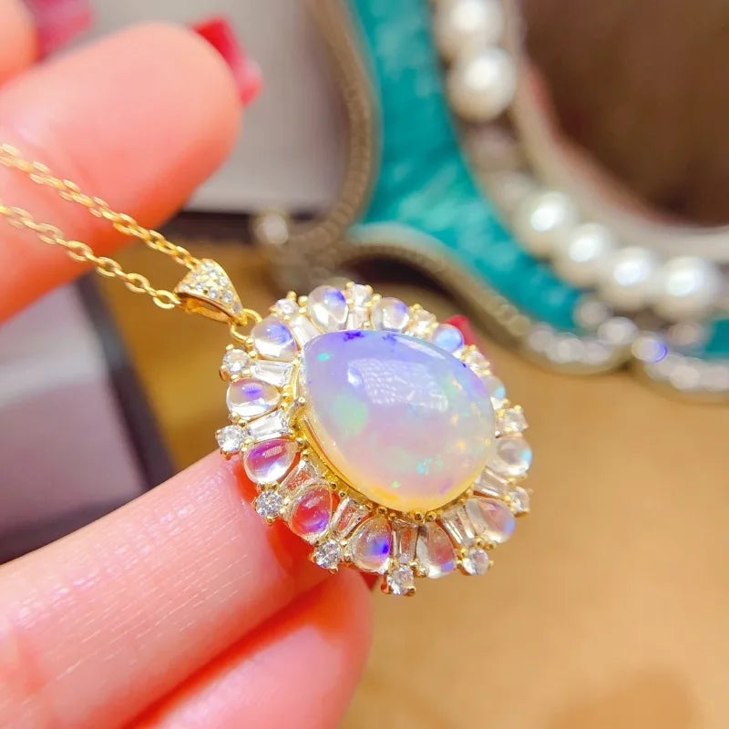 A gold pendant necklace with a large iridescent white opal center stone and small clear stones around it, held between fingers against a softly blurred jewelry background.