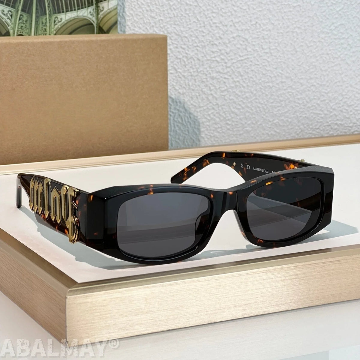A pair of designer sunglasses with a glossy black frame, dark lenses, and ornate gold-toned temple arms featuring the brand name 'Miu Miu', displayed on a light wood block.