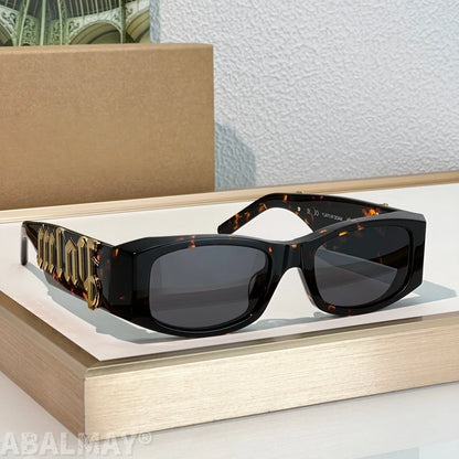 A pair of designer sunglasses with a glossy black frame, dark lenses, and ornate gold-toned temple arms featuring the brand name 'Miu Miu', displayed on a light wood block.