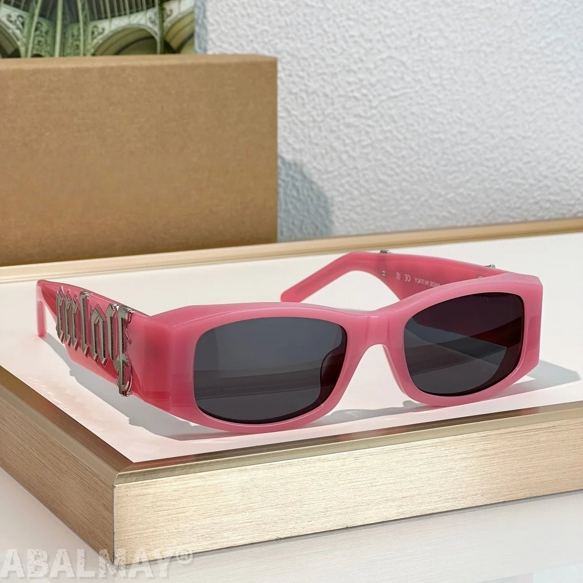 Pink rectangular sunglasses with dark lenses and a metallic gothic-style logo on the temple arm, displayed on a wooden tray with a cardboard box in the background.