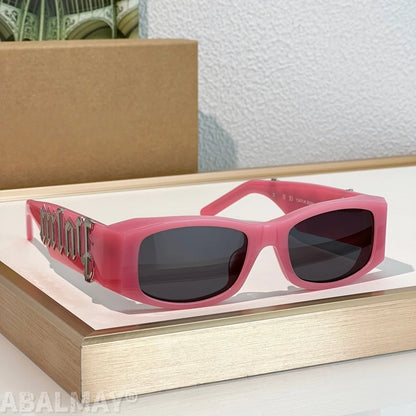 Pink rectangular sunglasses with dark lenses and a metallic gothic-style logo on the temple arm, displayed on a wooden tray with a cardboard box in the background.