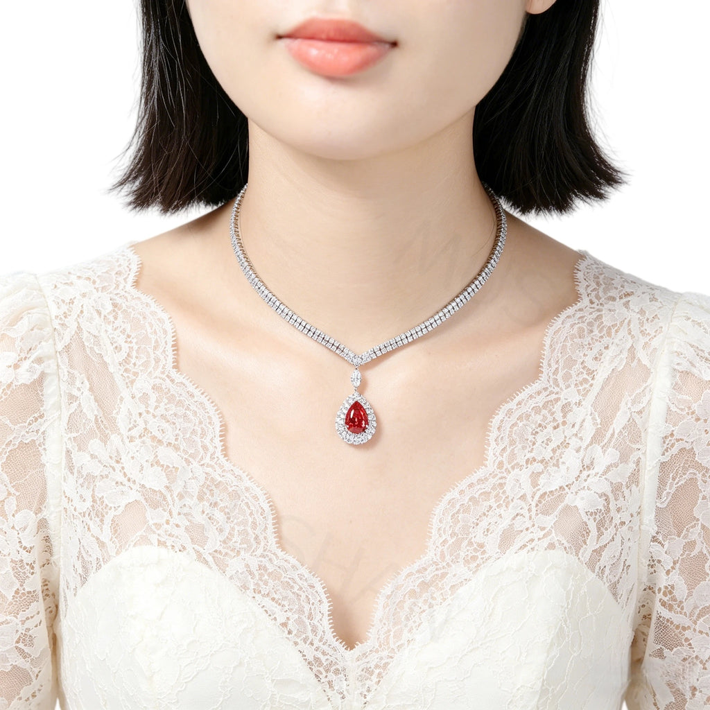 A close-up of a model wearing a silver-toned diamond-studded necklace with a teardrop-shaped red gemstone pendant, paired with a white lace top against a clean white background.