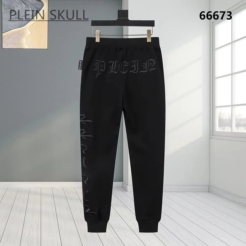 Black sweatpants with gothic-style embroidered branding and vertical text on the leg, hanging on a black hanger against a modern room background.