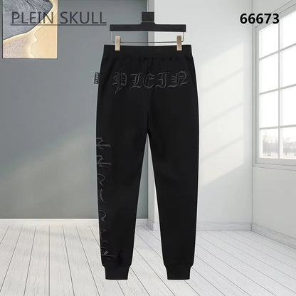Black sweatpants with gothic-style embroidered branding and vertical text on the leg, hanging on a black hanger against a modern room background.