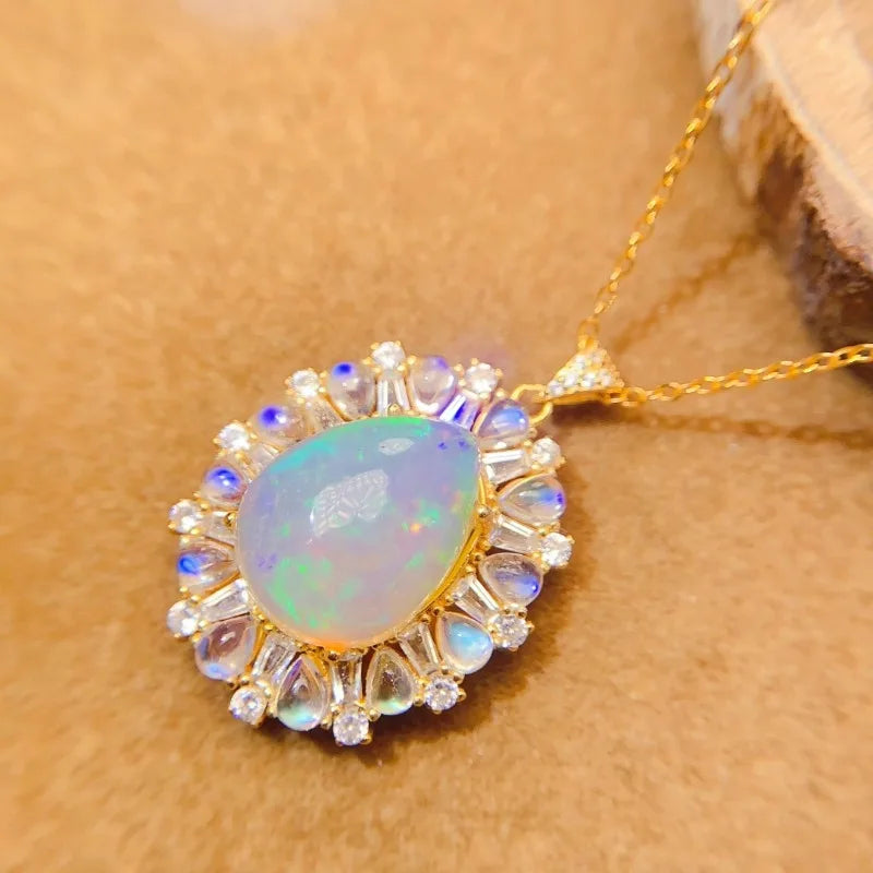 A gold pendant necklace featuring a teardrop-shaped iridescent opal stone surrounded by small clear stones and blue accent stones, displayed on a textured brown surface.