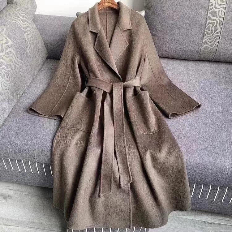 A taupe-brown wool coat with a matching waist tie belt, hanging on a wooden hanger on a gray fabric sofa.