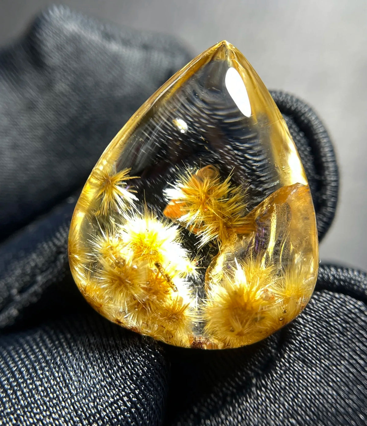 A teardrop-shaped clear resin cabochon containing yellow dried flower buds, resting on a textured black fabric surface.