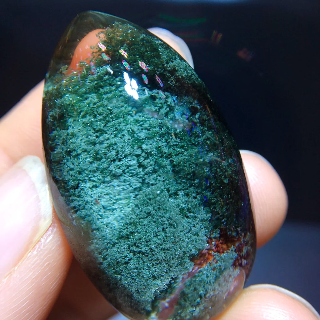 A polished dark green stone or crystal held between fingers, showing a textured mottled green surface with some iridescent blue and reddish reflections near the top.