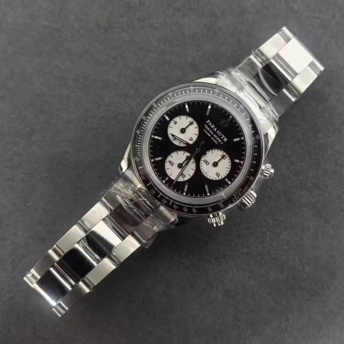 A stainless steel chronograph watch with a black dial and three white subdials, photographed diagonally on a dark gray textured background. The watch is still wrapped in protective plastic.