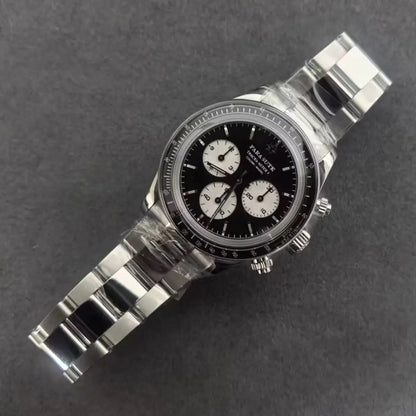 A stainless steel chronograph watch with a black dial and three white subdials, photographed diagonally on a dark gray textured background. The watch is still wrapped in protective plastic.