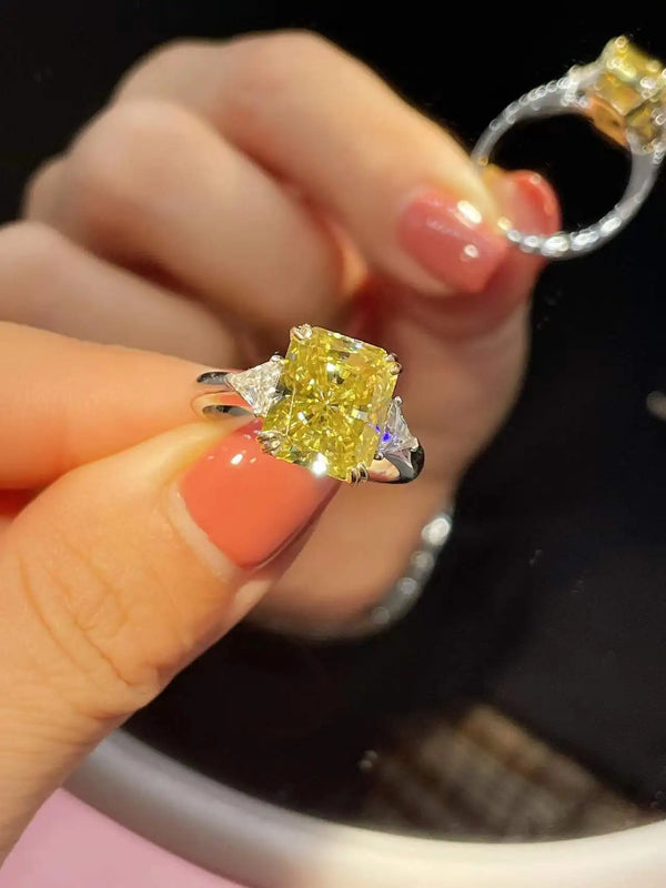 A close-up of a hand holding a yellow cushion-cut diamond ring with a silver-toned band, with another ring visible in the background against a dark surface.