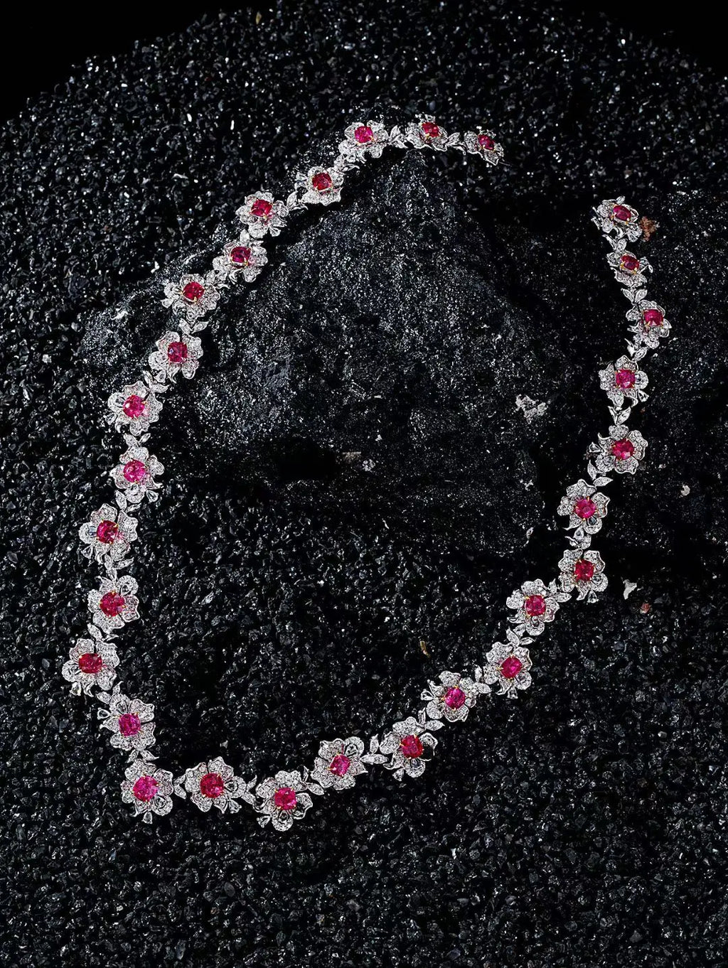 A silver-toned floral necklace with bright pink gemstone centers, arranged in an oval on a textured black background.