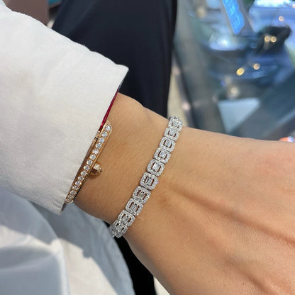 A close-up of a person's wrist wearing two bracelets: a silver-toned square-link bracelet paved with clear stones and a gold-toned bangle with a single clear stone, shown against a blurred background.