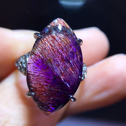 A close-up of a silver ring featuring a large marquise-shaped purple iridescent gemstone with fine metallic needle-like inclusions, held on a person's fingers against a dark background.