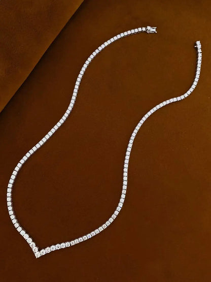 A single-strand diamond tennis necklace in a silver-toned setting, arranged in a curved S-shape on a textured brown background.