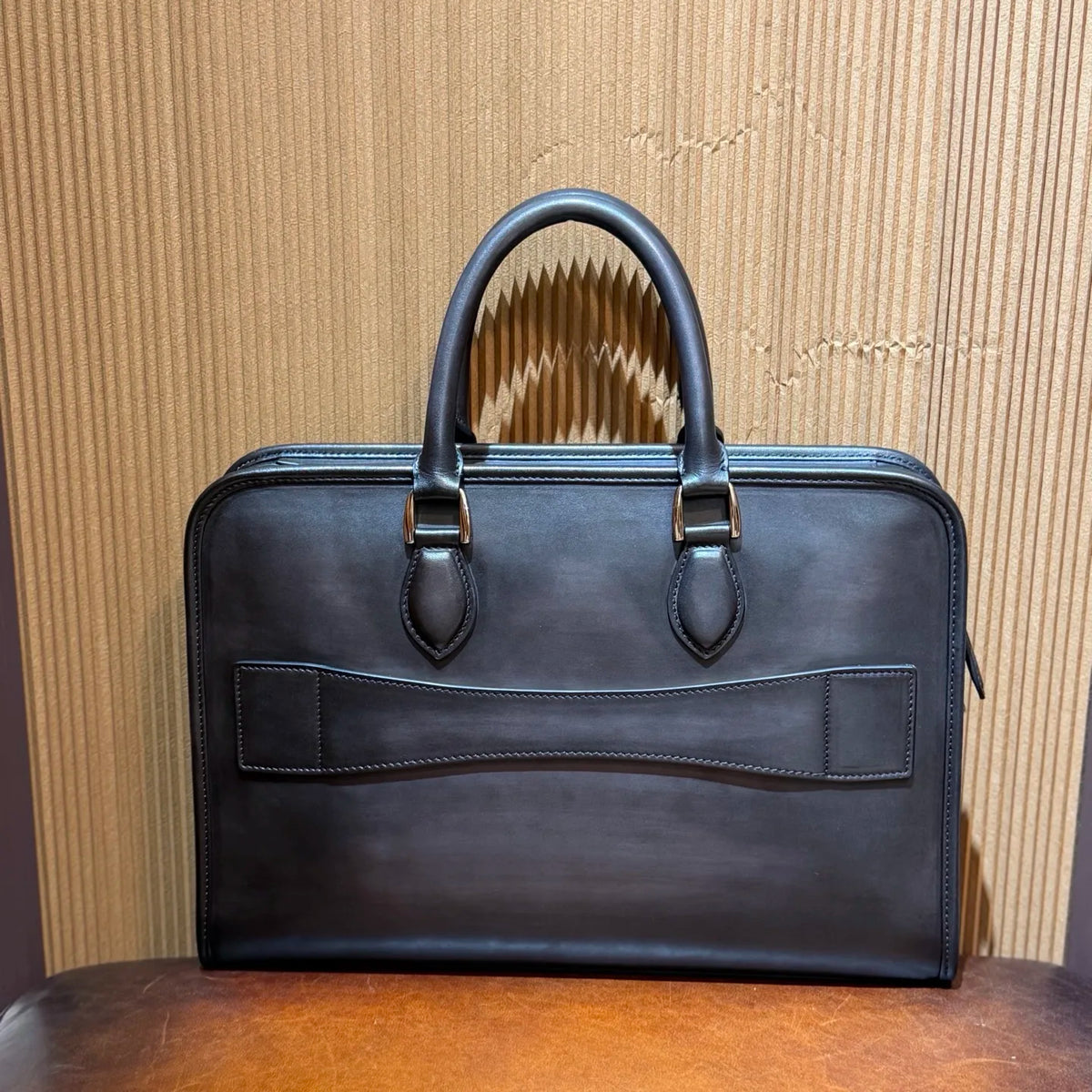 A dark brown leather briefcase with double top handles and brass hardware, displayed on a wooden surface against a corrugated cardboard background.